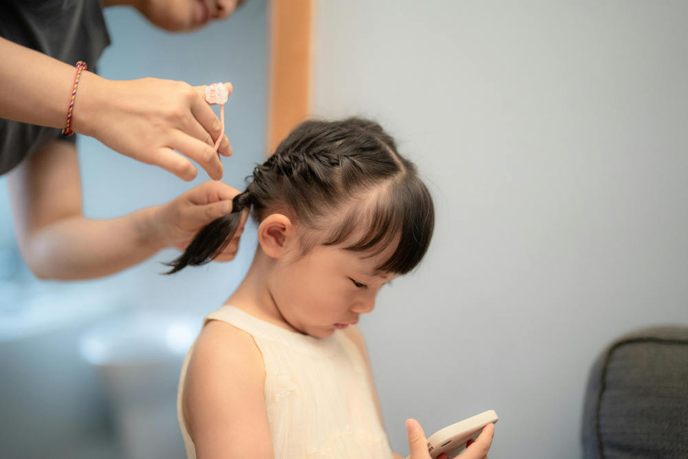 A woman is gently doing a young girl's hair at home, emphasizing a loving and attentive caregiving moment.